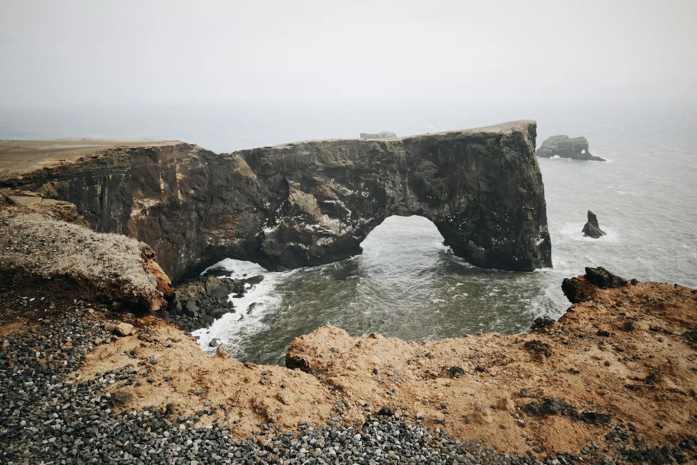 Large natural rock arch over the sea seen from a gravel clifftop, small sea stacks in hazy light