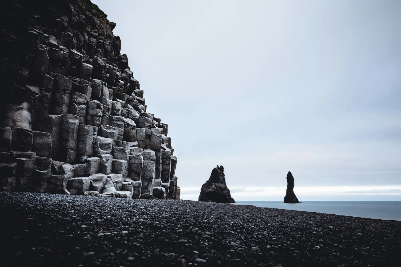 Wall of hexagonal basalt columns beside a black pebble beach, two sea stacks offshore