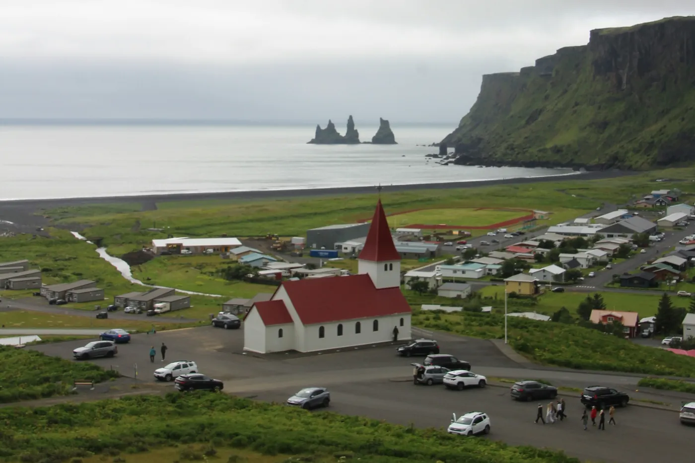 White church with red roof above the village of Vík, green cliffs and the Reynisdrangar stacks on the coast