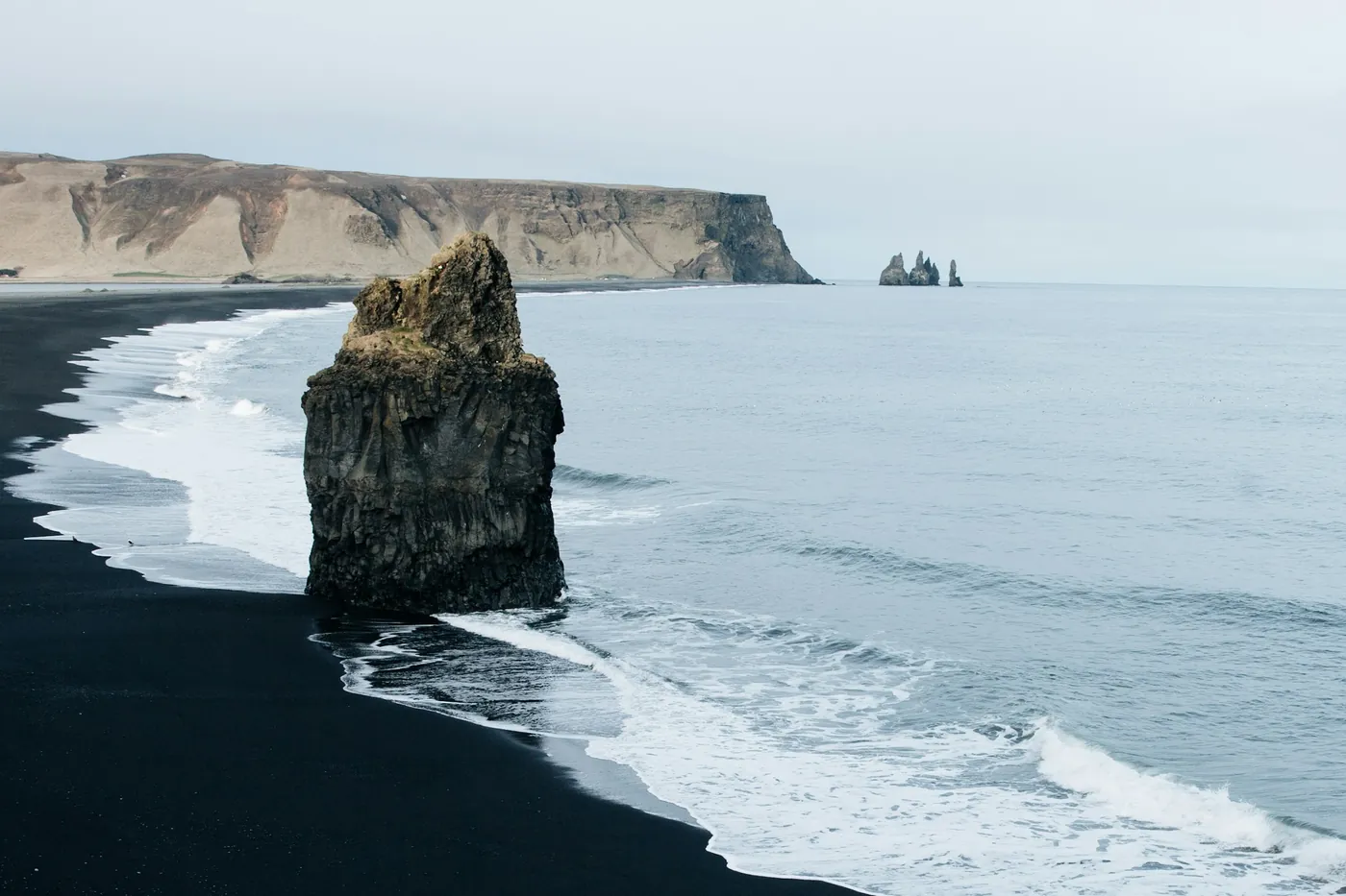 Lone basalt sea stack on a black sand beach with surf, long headland and distant sea stacks behind