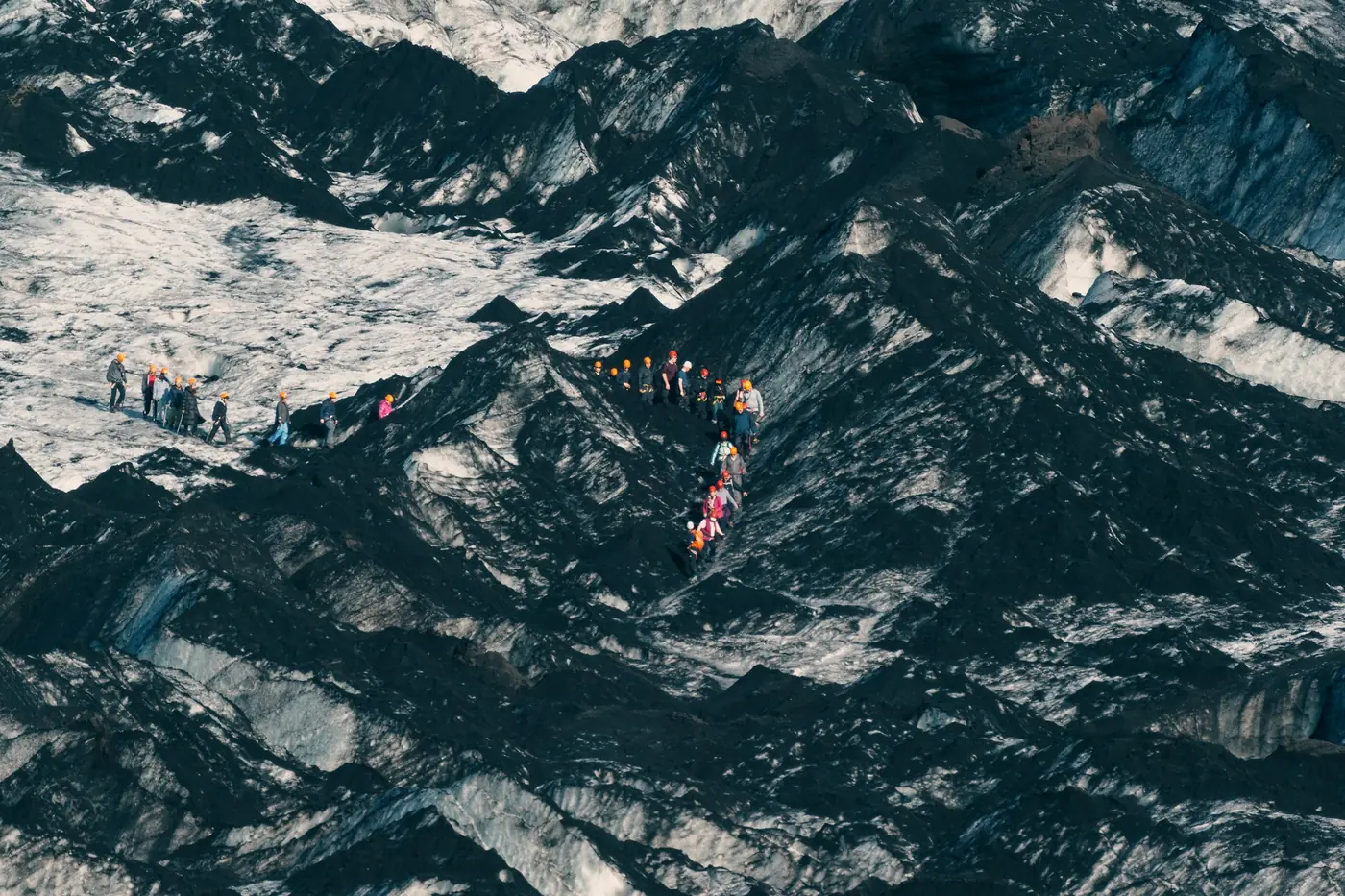 Aerial view of a group of hikers in a line crossing a glacier streaked with black ash