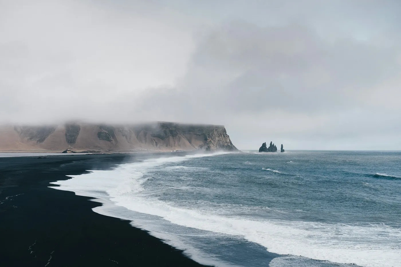 Waves rolling onto a curved black sand beach, misty headland with the Reynisdrangar sea stacks offshore