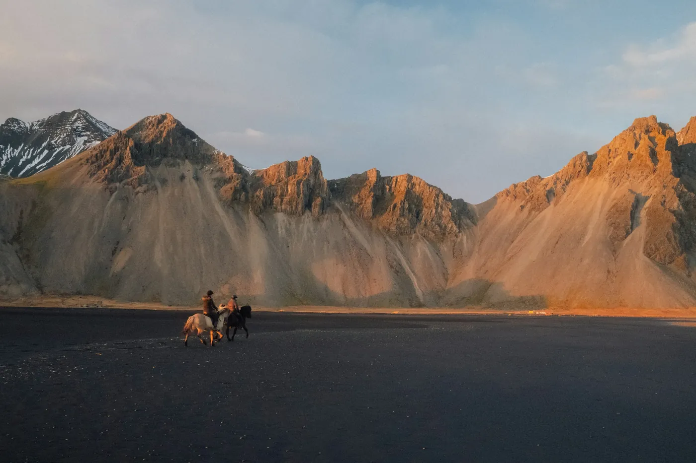 Two riders on Icelandic horses crossing a black sand plain below jagged mountains in evening light