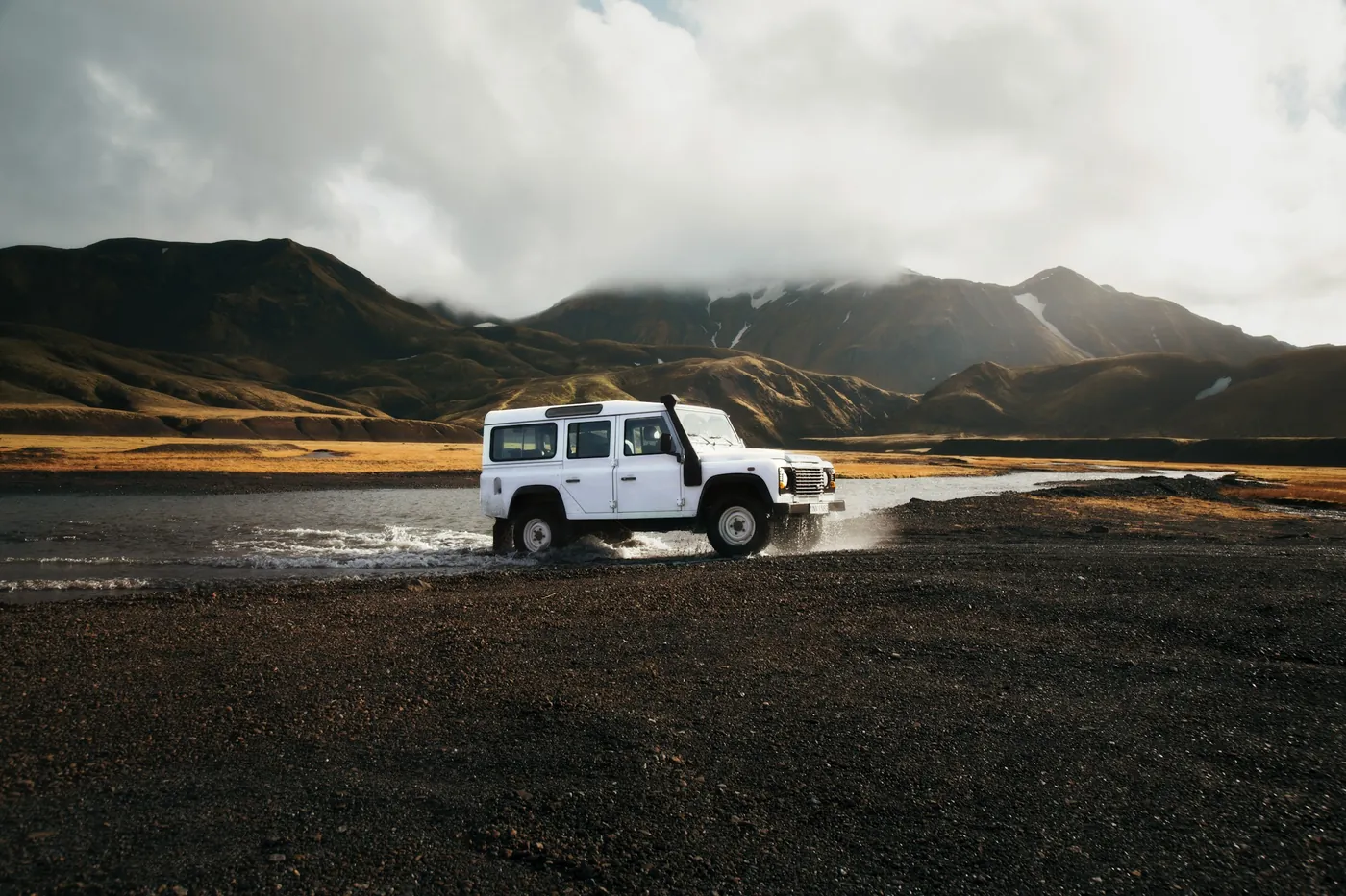 White off-road vehicle splashing through a shallow river on a gravel plain, mountains behind