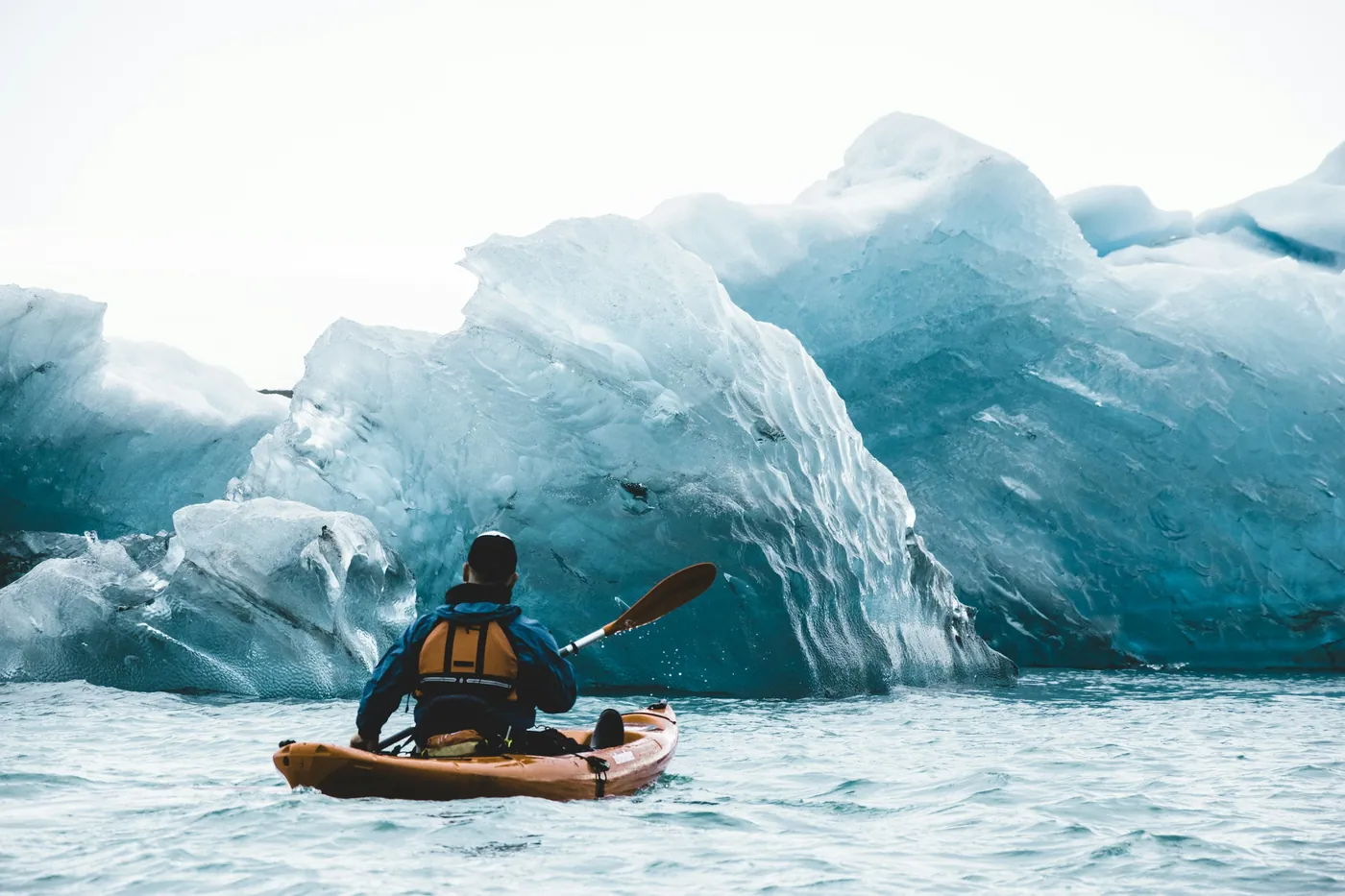 Kayaker in an orange boat seen from behind, paddling towards large blue icebergs