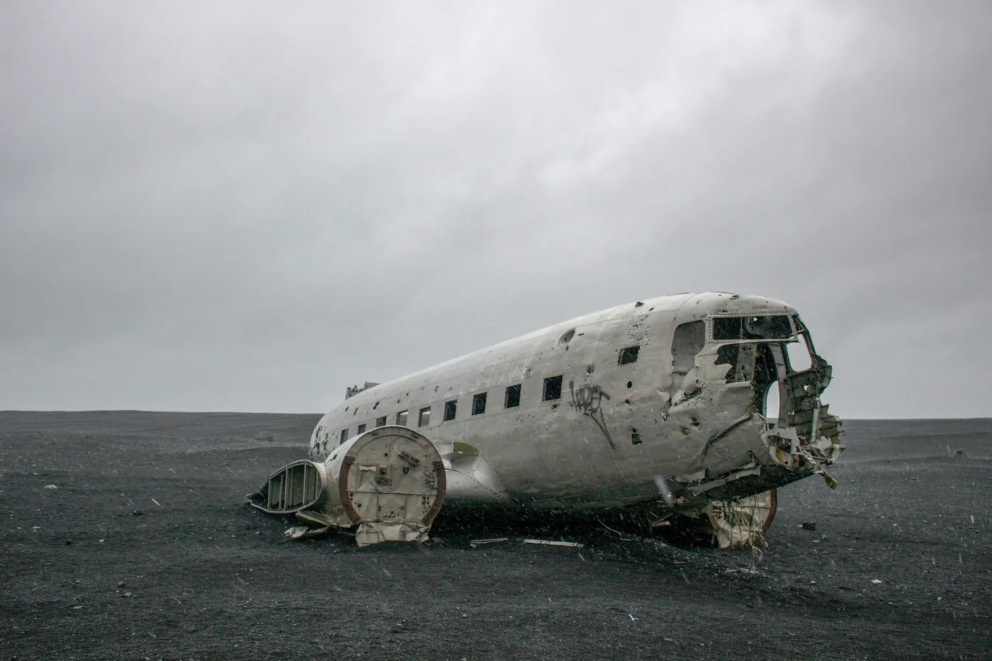 Fuselage of the wrecked DC-3 on a flat black sand plain under a grey sky