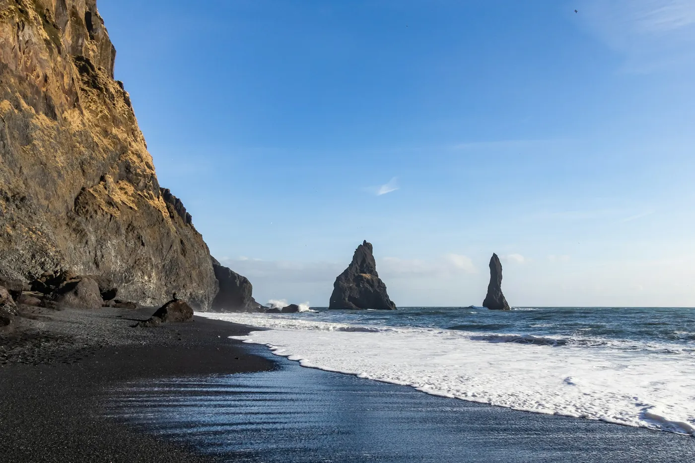 Basalt sea stacks rising from the surf off a black sand beach below a sunlit cliff