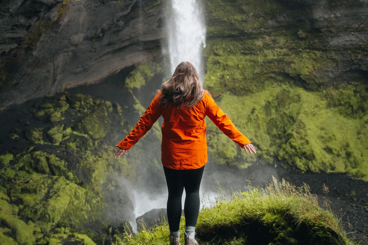 Woman in an orange rain jacket seen from behind, arms out, in front of a waterfall in a mossy gorge